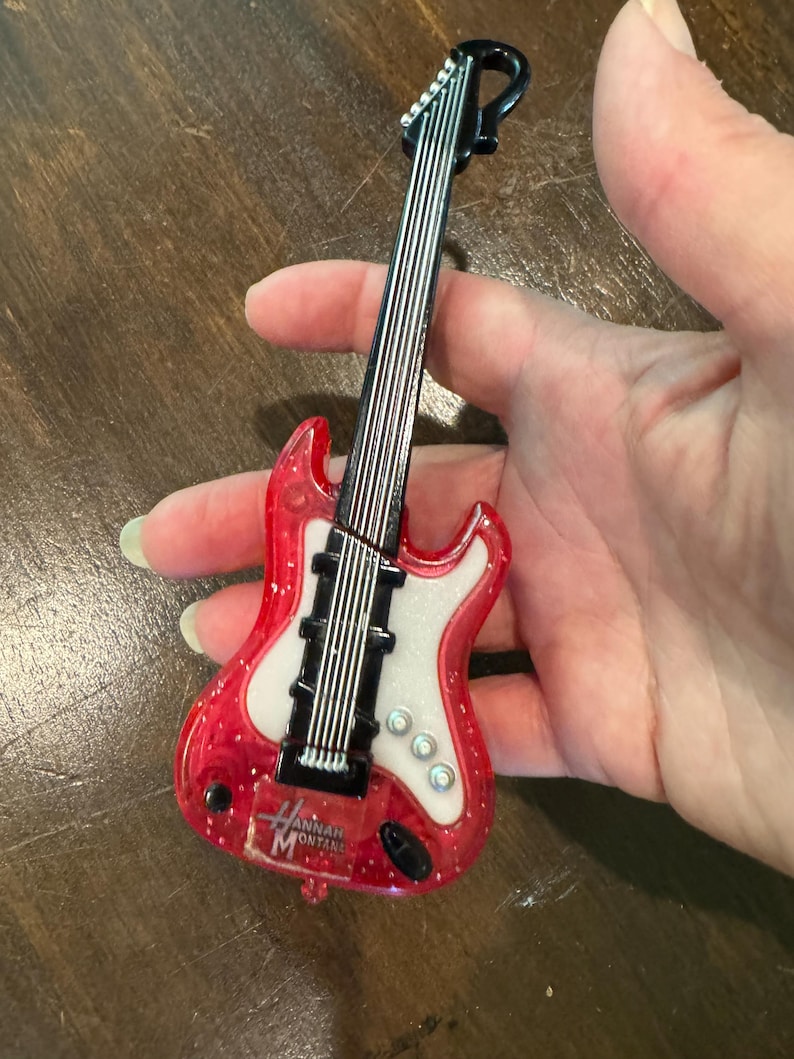 Puede incluir: Una guitarra roja y blanca en miniatura con el texto "Hannah Montana". La guitarra tiene cuerdas negras y un clavijero negro. El cuerpo de la guitarra es rojo con purpurina y un centro blanco.