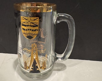 May include: A clear glass beer mug with a gold rim and a black and gold design featuring the Lions Gate Bridge and the coat of arms of the city of Vancouver, British Columbia. The text "Lions Gate Bridge" is printed on the mug.