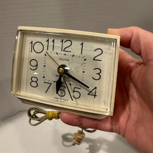 May include: A white dialite electric alarm clock with black numbers and hands. The clock has a square face and is plugged in with a cord.