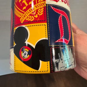May include: A cylindrical Disney-themed mug with a patchwork design. The mug features the Disneyland Resort logo, the letter "D", and a Mickey Mouse silhouette. The color scheme includes red, yellow, blue, and white.