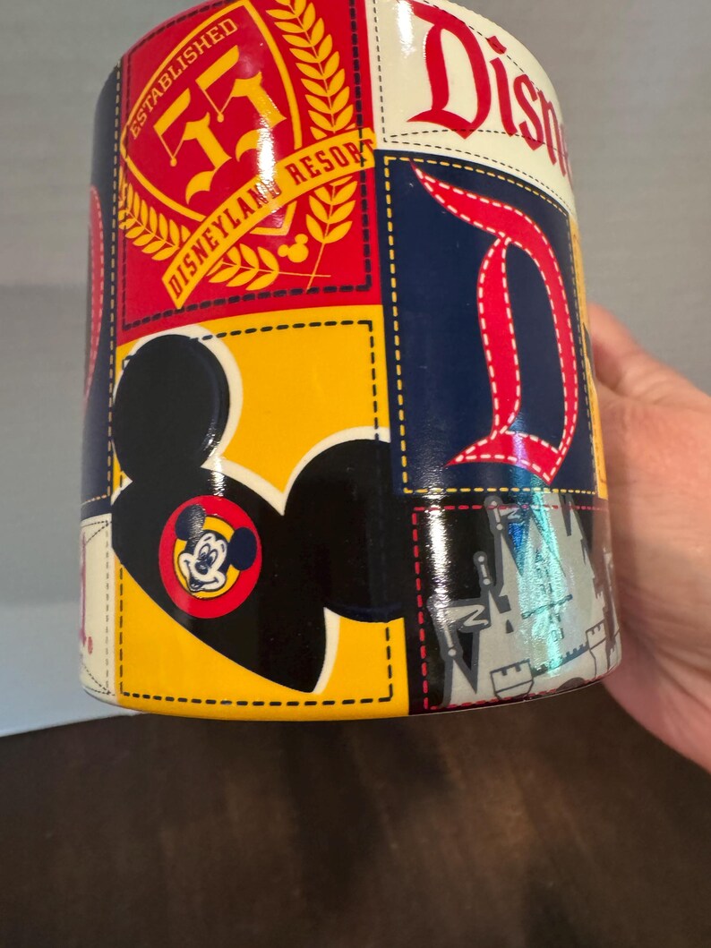 May include: A cylindrical Disney-themed mug with a patchwork design. The mug features the Disneyland Resort logo, the letter "D", and a Mickey Mouse silhouette. The color scheme includes red, yellow, blue, and white.