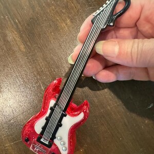 Puede incluir: Un adorno en miniatura con forma de guitarra el&eacute;ctrica roja y blanca, con cuerdas negras y un clavijero negro. El adorno tiene el texto "Hannah Montana" en el cuerpo. Es un art&iacute;culo de novedad.