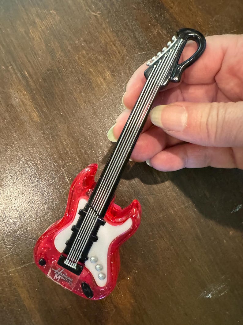 Puede incluir: Un adorno en miniatura con forma de guitarra el&eacute;ctrica roja y blanca, con cuerdas negras y un clavijero negro. El adorno tiene el texto "Hannah Montana" en el cuerpo. Es un art&iacute;culo de novedad.