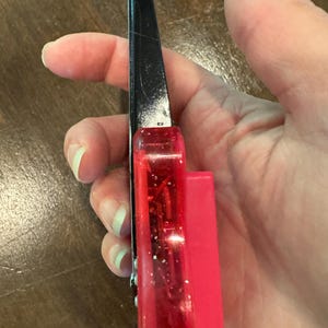 Puede incluir: Un corta&uacute;&ntilde;as negro y rojo con un mango de pl&aacute;stico rojo transparente y un accesorio de pl&aacute;stico rosa brillante. El corta&uacute;&ntilde;as se sostiene en una mano, mostrando la vista lateral de la herramienta.