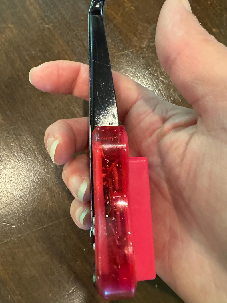 Puede incluir: Un corta&uacute;&ntilde;as negro y rojo con un mango de pl&aacute;stico rojo transparente y un accesorio de pl&aacute;stico rosa brillante. El corta&uacute;&ntilde;as se sostiene en una mano, mostrando la vista lateral de la herramienta.