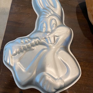 May include: A silver-colored Bugs Bunny-shaped cake pan. The pan is made of metal and features the iconic cartoon character's face, ears, and body. The pan is designed to bake a cake in the shape of Bugs Bunny.