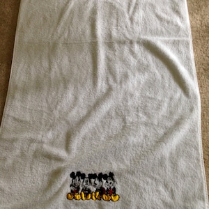 Puede incluir: Toalla de ba&ntilde;o blanca con una imagen bordada de cuatro personajes de Mickey Mouse en fila. Los personajes llevan pantalones cortos rojos y zapatos amarillos.