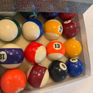 May include: A set of billiard balls in a box. The balls are a mix of colors, including yellow, red, blue, and black, each with a white stripe and a number. The balls are arranged neatly inside the box.