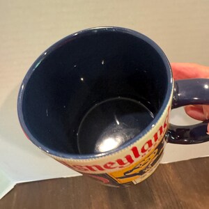 May include: A blue ceramic mug with a handle, featuring the word "Disneyland" in red and yellow lettering. The interior of the mug is a deep blue, and the handle is also blue.