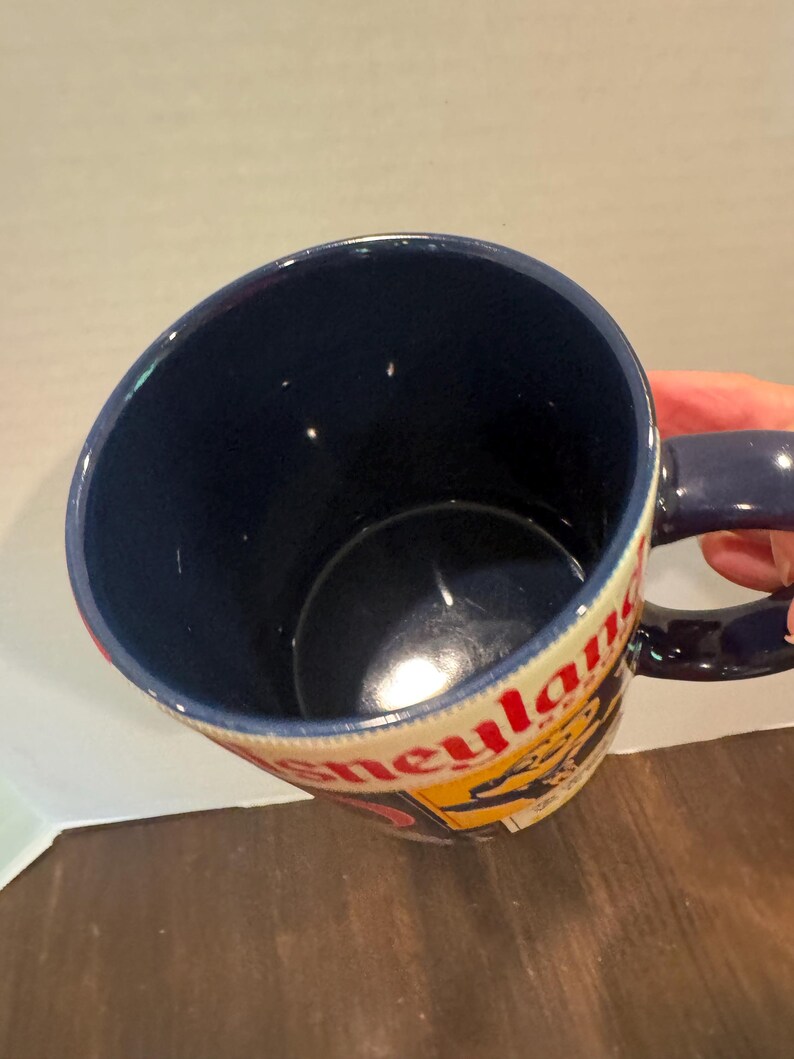 May include: A blue ceramic mug with a handle, featuring the word "Disneyland" in red and yellow lettering. The interior of the mug is a deep blue, and the handle is also blue.