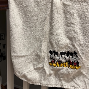 Puede incluir: Toalla de ba&ntilde;o blanca con un dise&ntilde;o bordado de cuatro personajes de Mickey Mouse en fila. Los personajes llevan pantalones cortos rojos y zapatos amarillos.