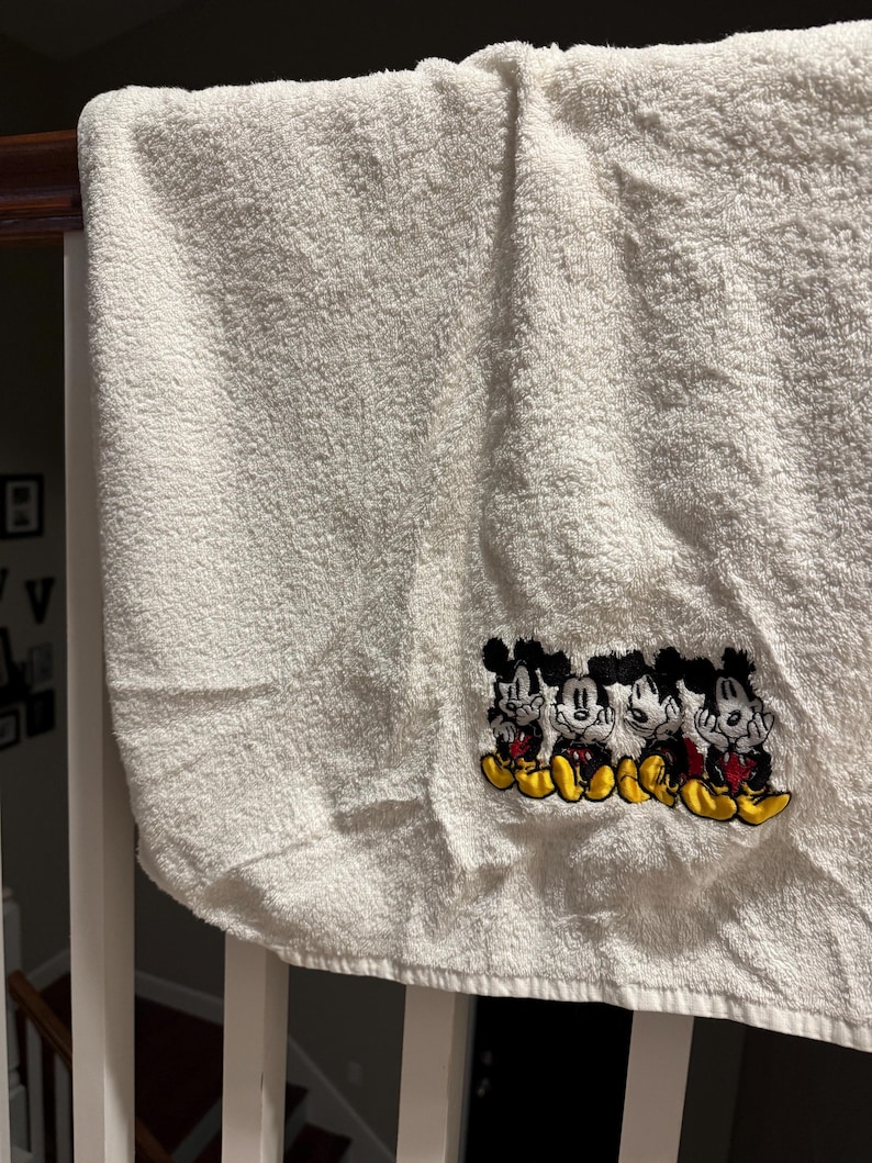 Puede incluir: Toalla de ba&ntilde;o blanca con un dise&ntilde;o bordado de cuatro personajes de Mickey Mouse en fila. Los personajes llevan pantalones cortos rojos y zapatos amarillos.