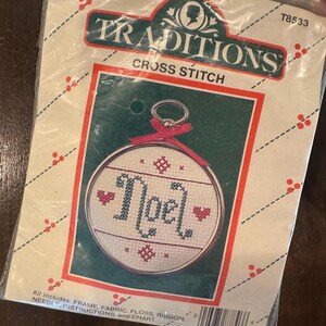 May include: A Traditions cross stitch kit featuring a round design with the word "Noel" in blue cross-stitch, accented with red hearts and stars. The kit includes a frame, fabric, floss, ribbon, needle, instructions, and chart. The packaging is cream-colored with red and green accents.