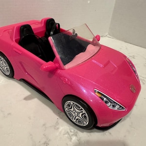 May include: A bright pink toy convertible car with black seats and silver-colored wheels. The car has a clear windshield and side mirrors. The car's body has a metallic sheen.