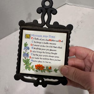 May include: Black cast iron trivet with a decorative frame and handle. The trivet features a ceramic tile with a poem titled "Mother and Dad" with floral illustrations in blue, orange, and green.