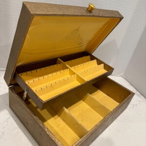 May include: A vintage two-tiered jewelry box with a gold-toned exterior and yellow velvet lining. The top tier has multiple compartments for storing rings, earrings, and other small jewelry items. The bottom tier has a larger compartment for storing necklaces, bracelets, or other larger jewelry items.