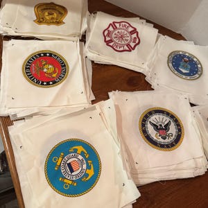 May include: A stack of white fabric squares with different embroidered patches. The patches feature the logos of the United States Marine Corps, United States Coast Guard, United States Navy, and a fire department.