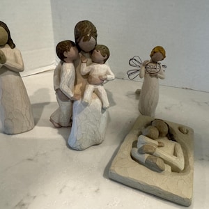 May include: A collection of four cream-colored resin figurines featuring a standing figure with hands together, a central figure holding two children, an angel with wire wings holding a circle marked 2007, and a rectangular plaque with a relief.