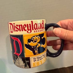 May include: A Disneyland Resort coffee mug with a navy blue handle. The mug features a patchwork design with the Disneyland logo in red, a Mickey Mouse graphic, and the text "The Happiest Place on Earth".