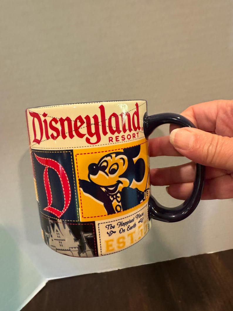 May include: A Disneyland Resort coffee mug with a navy blue handle. The mug features a patchwork design with the Disneyland logo in red, a Mickey Mouse graphic, and the text "The Happiest Place on Earth".