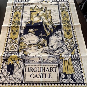 May include: Cream-colored Urquhart Castle tea towel with a detailed illustration in black, gold, and navy. The design features the castle, figures, and the text "Urquhart Castle". The towel is made of 100% cotton.