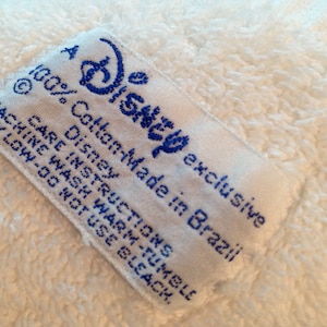 Puede incluir: Toalla de algod&oacute;n blanca con una etiqueta azul y blanca que dice "A Disney exclusive 100% Cotton-Made in Brazil Disney Care Instructions Machine Wash Warm-Tumble Dry Low-Do Not Use Bleach."