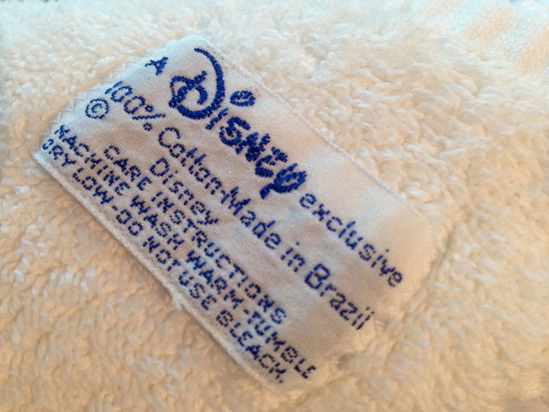Puede incluir: Toalla de algod&oacute;n blanca con una etiqueta azul y blanca que dice "A Disney exclusive 100% Cotton-Made in Brazil Disney Care Instructions Machine Wash Warm-Tumble Dry Low-Do Not Use Bleach."