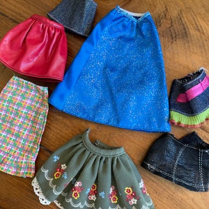 May include: A collection of six doll skirts in various colors and patterns, including red, blue, green, black, and pink. The skirts are made of different fabrics, such as denim, corduroy, and plaid.