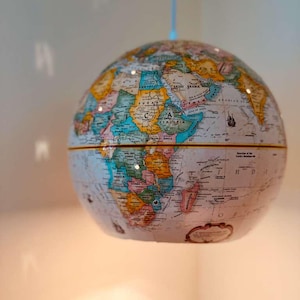Könnte beinhalten: Ein Globus im Vintage-Stil mit einer detaillierten Weltkarte. Der Globus ist in zwei Hälften geteilt und zeigt Kontinente und Länder in verschiedenen Farben. Er hängt an einer dünnen, hellblauen Schnur, was auf einen dekorativen Zweck hindeutet.