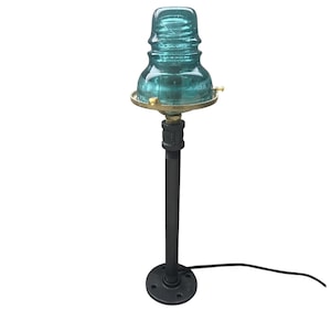 May include: A table lamp featuring a translucent teal glass insulator as the shade, mounted on a black metal pipe stand with a circular base. The insulator is secured by a brass-colored metal ring with small screws, and a black power cord extends.
