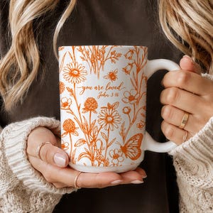May include: A white ceramic mug featuring an orange botanical print of wildflowers, a butterfly, and a bee. The text "you are loved" and "John 3:16" is printed in orange script and serif font on the front of the mug, which has a curved handle.