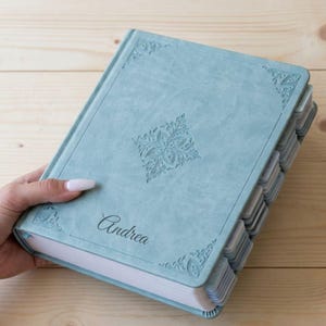 Może przedstawiać: Jasnoniebieski notatnik z wytłoczonym imieniem "Andrea" na okładce. Notatnik posiada dekoracyjne wzory w rogach i centralny kwiatowy wzór. Strony są podzielone na zakładki.