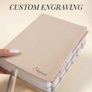 Op de afbeelding: Een beige boek met de tekst "CUSTOM ENGRAVING" bovenaan. Het boek heeft een gepersonaliseerde naam, "Stephanie", gegraveerd op de kaft. Het boek heeft meerdere gekleurde paginascheiders en twee lint-bladwijzers.