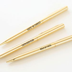 May include: Three gold-coloured pens with black text. The pens are arranged diagonally, with the text reading "HOPE IN CHRIST", "FAITH OVER FEAR", and "WALK BY FAITH". The pens have a sleek, minimalist design.