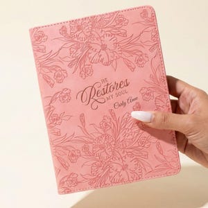 Op de afbeelding: Een roze notitieboek met bloemreliëf. De kaft bevat de tekst "He Restores My Soul" en de naam "Carly Anne" in een sierlijk lettertype. Het notitieboek wordt vastgehouden door een hand tegen een lichte achtergrond.