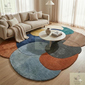 May include: A colourful, abstract circular rug with sections of blue, orange, brown, and grey. A beige sofa with cushions and a throw blanket is in the background. A round coffee table sits on the rug, with a vase and books.