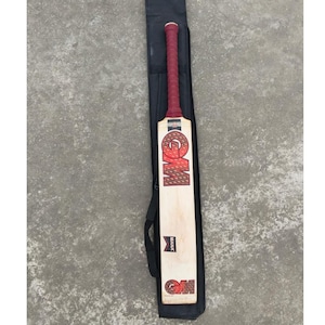 May include: A cricket bat with a red grip and a light-colored wooden blade. The bat is in a black carrying case. The bat has the letters "GM" in a red and white design on the blade. The word "MANA" is also visible.