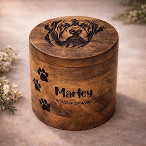 May include: A cylindrical wooden pet urn featuring a carved dog portrait on the lid. The urn is engraved with paw prints, the name "Marley", and dates 19/03/2010-21/04/2025. The wood has a natural grain.