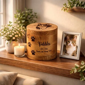 Peut inclure: Urne funéraire en bois avec un portrait de chat gravé sur le couvercle et des empreintes de pattes sur le côté. Le nom "Pebbles", les dates 2011-2026 et la phrase "forever and always loved" sont gravés. Une photo encadrée d'un chat est à côté.