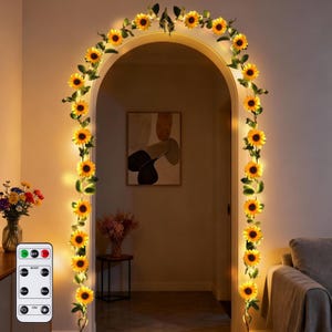 May include: A decorative archway adorned with a sunflower garland and warm white LED lights. A remote control is visible on the left. The garland is wrapped around the archway and doorway, creating a welcoming and festive atmosphere.