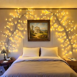 May include: A bedroom scene featuring a bed with white pillows and a neutral-colored comforter. The wall is adorned with string lights in a tree branch design, and a framed painting hangs above the bed. A lamp sits on a side table.