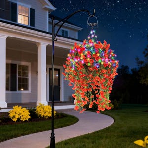 May include: A black metal hanging basket stand with a curved arm supporting a hanging basket filled with red flowers. The flowers are illuminated with colorful LED lights, creating a vibrant display. The scene is set at night with a house in the background.