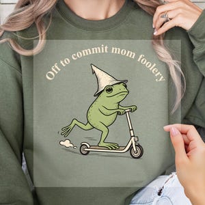 May include: An olive green crewneck sweatshirt featuring a graphic of a green frog wearing a pointed hat and riding a kick scooter. The text above the frog reads, "Off to commit mom foolery" in a light-colored, curved, sans-serif font.