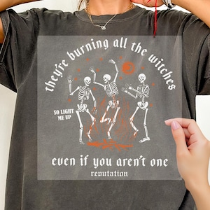 May include: A charcoal gray t-shirt featuring a graphic print of three skeletons dancing around a fire. The text reads, they're burning all the witches, so light me up, even if you aren't one, and reputation in a white gothic-style font.