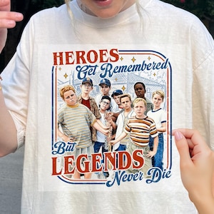 May include: Cream-colored t-shirt featuring a graphic of a group of boys with the text "HEROES Get Remembered But LEGENDS Never Die" in red and blue. The graphic is framed with a blue border and stars.