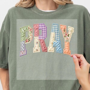 May include: An olive green crew neck t-shirt featuring the word PRAY in large, block-style letters. Each letter is designed with a patchwork aesthetic, incorporating various patterns including floral prints, gingham checks, and polka dots in pastel tones.
