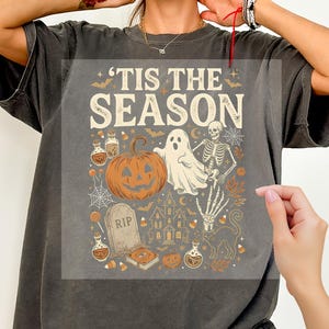 May include: A charcoal gray t-shirt featuring a graphic print with the text 'TIS THE SEASON. The design includes a jack-o'-lantern, a ghost, a skeleton, a tombstone marked RIP, a haunted house, a black cat, potion bottles, and candy corn.