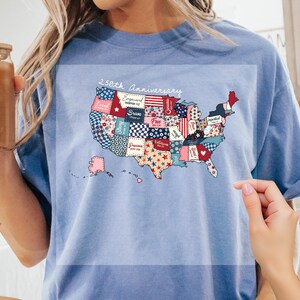 May include: Light blue t-shirt featuring a patchwork map of the United States with the text "250th Anniversary." The map includes various patriotic patterns and words like "Brave" and "Free."