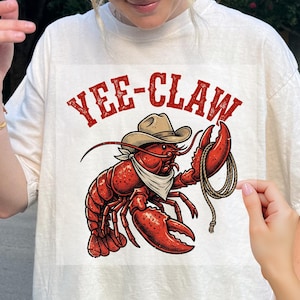 May include: White t-shirt featuring a red lobster wearing a cowboy hat and bandana, holding a rope. The text "YEE-CLAW" is printed above the lobster in a distressed red font. A fun, western-themed graphic.