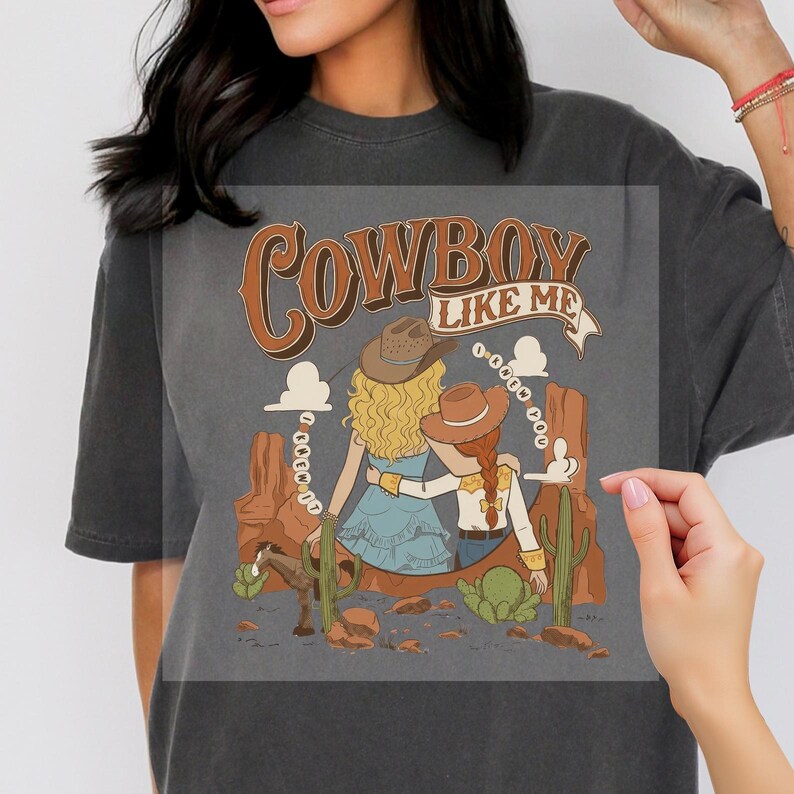 Pu&ograve; includere: Una t-shirt grigio antracite con una stampa grafica di due figure con cappelli da cowboy in un paesaggio desertico con cactus e un cavallo. Il testo "COWBOY LIKE ME" appare in alto, con "I KNEW IT" e "I KNEW YOU" curvati attorno.